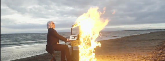 A person playing a burning piano at a sandy beach under a cloudy sky