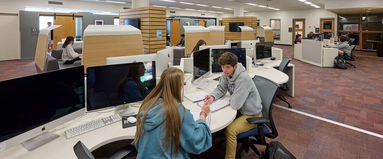  Two students talking to each other at the computers on the first floor of the library.