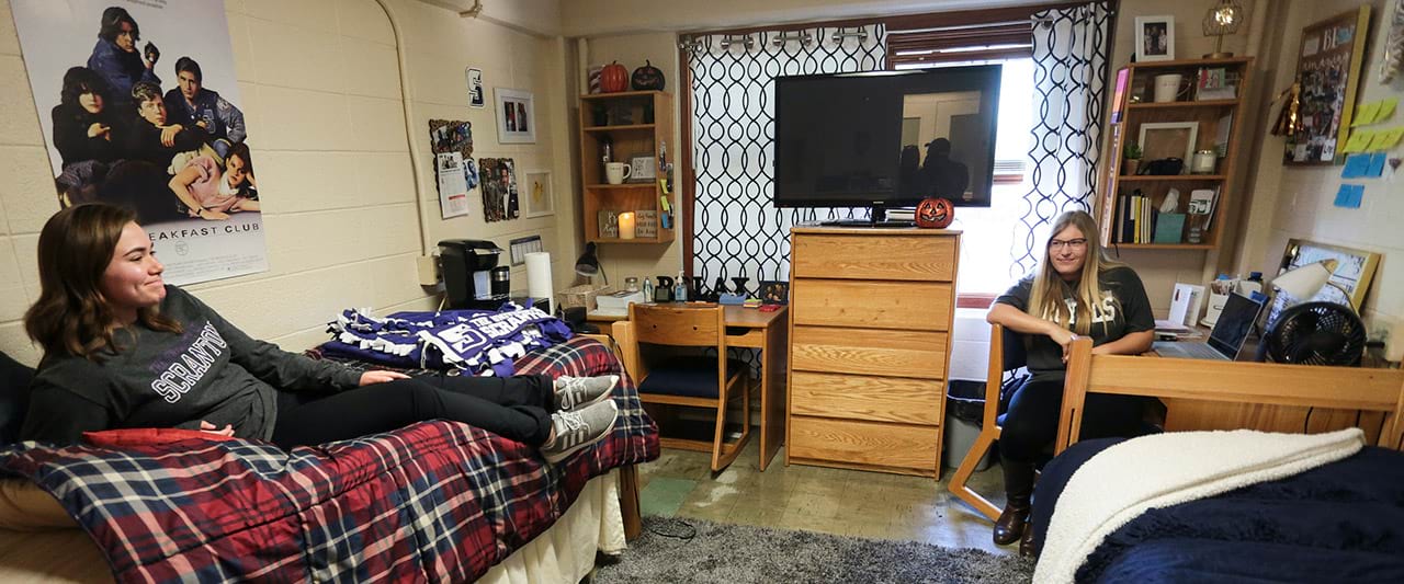  Two female roommates looking at each other sitting on their bed and desk chair in their  decorated dorm room. 