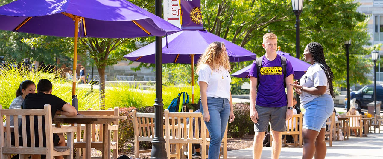  Three students talking to each other by the tables and umbrellas on the commons.