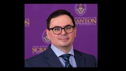 Dr. Ovidiu Cocieru in blue suit and tie against University purple background. 