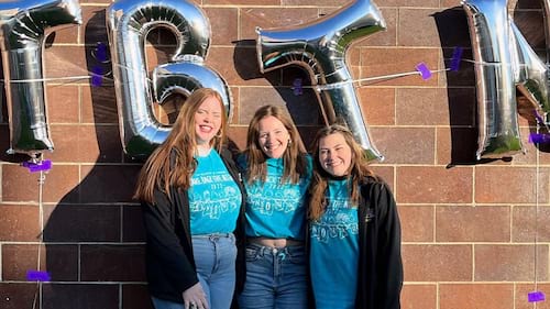 three women wearing purple T-shirts standing in front of large silver balloon letters T-B-T-N