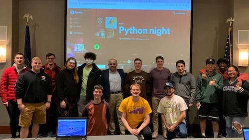 Shown are students and faculty assembled in a classroom. Behind them a whiteboard featuring the phrase "Python Night" 